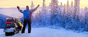 man stands next to his snowmobile with his arms outstretched towards the setting sun in front of snow covered trees