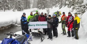 a group of 11 people in winter gear stand in front of a snowmobile holding a sign that says merry Christmas on the side of a snowmobile trail