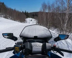first-person view of a snowmobile traveling down a snow-covered road between barren seasonal trees