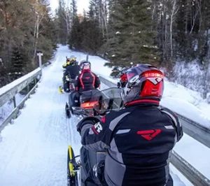 A line of snowmobilers cross a wooden bridge in the middle of a pine forest while wearing helmet safety lights