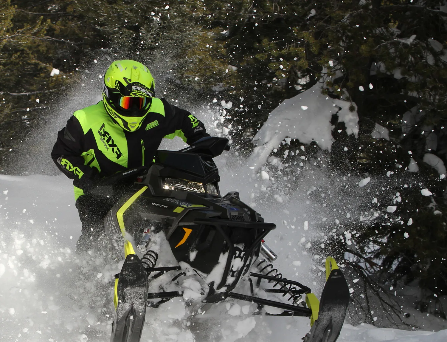 a man in modern green snowmobile gear plows through a snowbank in front of pine trees