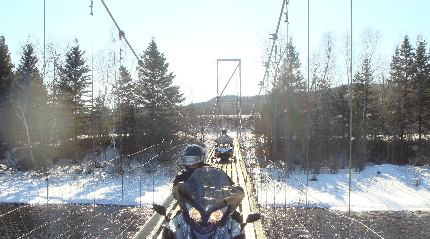 two snowmobilers cross a suspension bridge over a river
