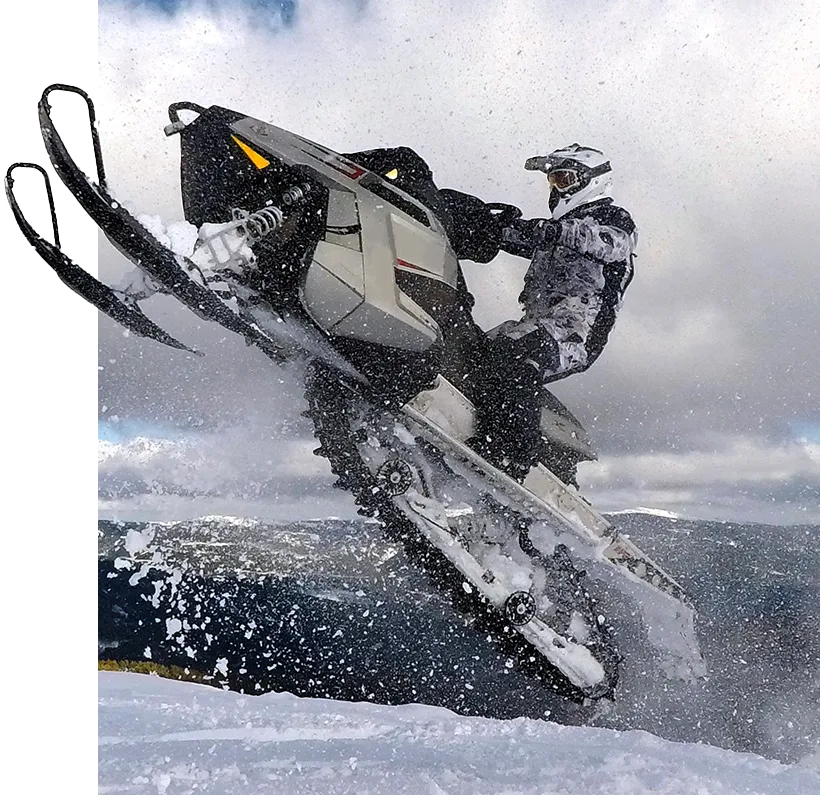 a snowmobiler just leaving the top of a jump with mountains in the background