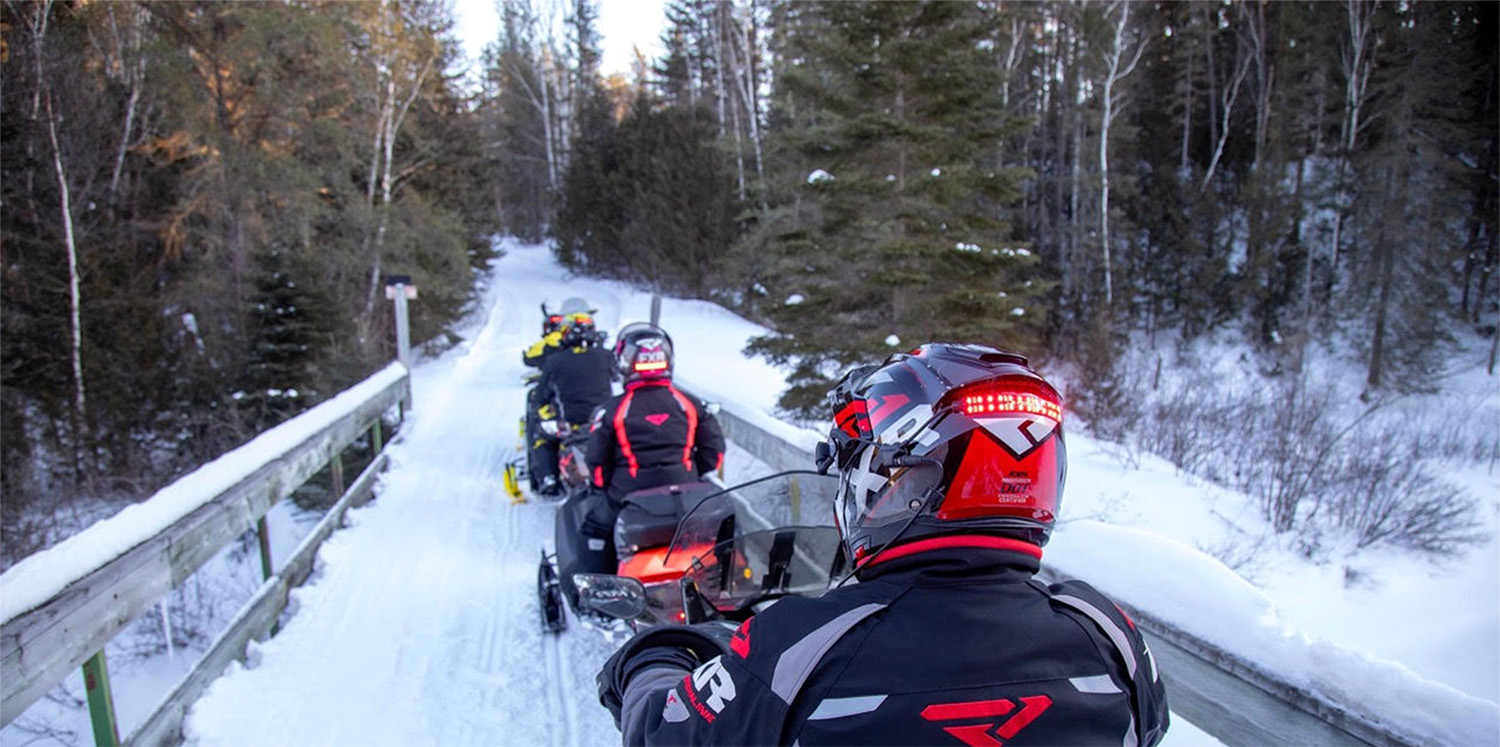 A group of three snowmobilers cross a snow-covered bridge on a path leading through a forest