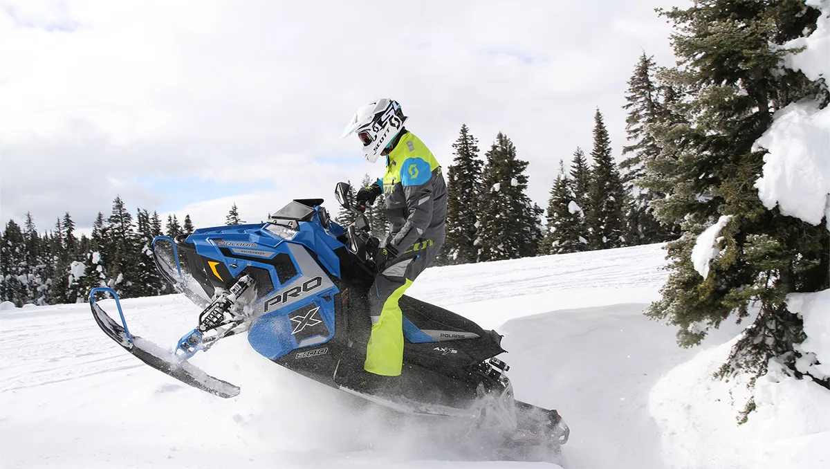 snowmobiler going off a jump in a large field covered in snow with snow-covered pine trees in the background