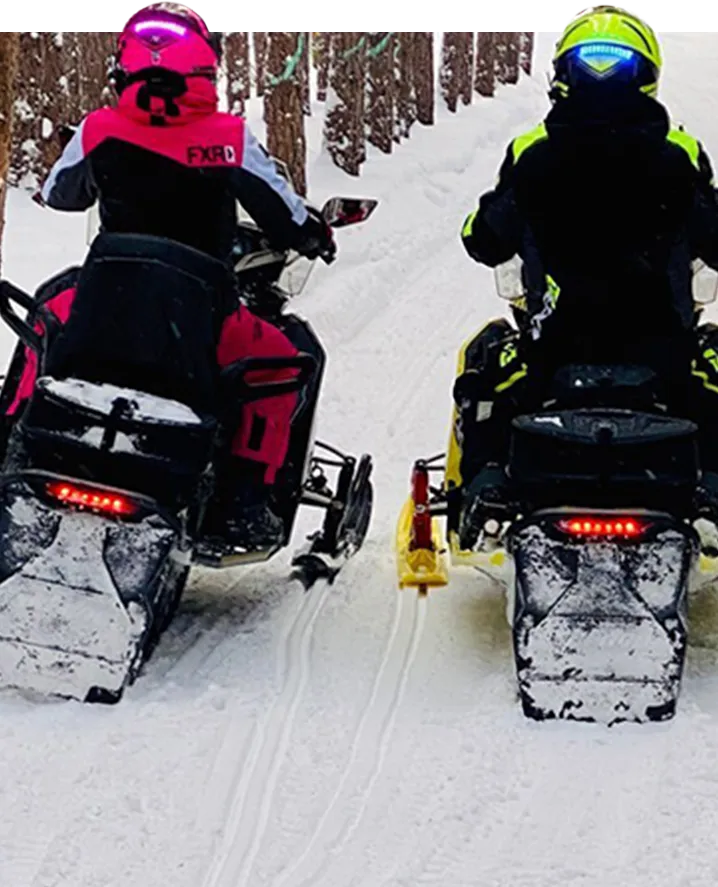 A man and woman side by side on their snowmobiles facing away from the camera showing a blue and pink helmet safety light on the back of their helmets